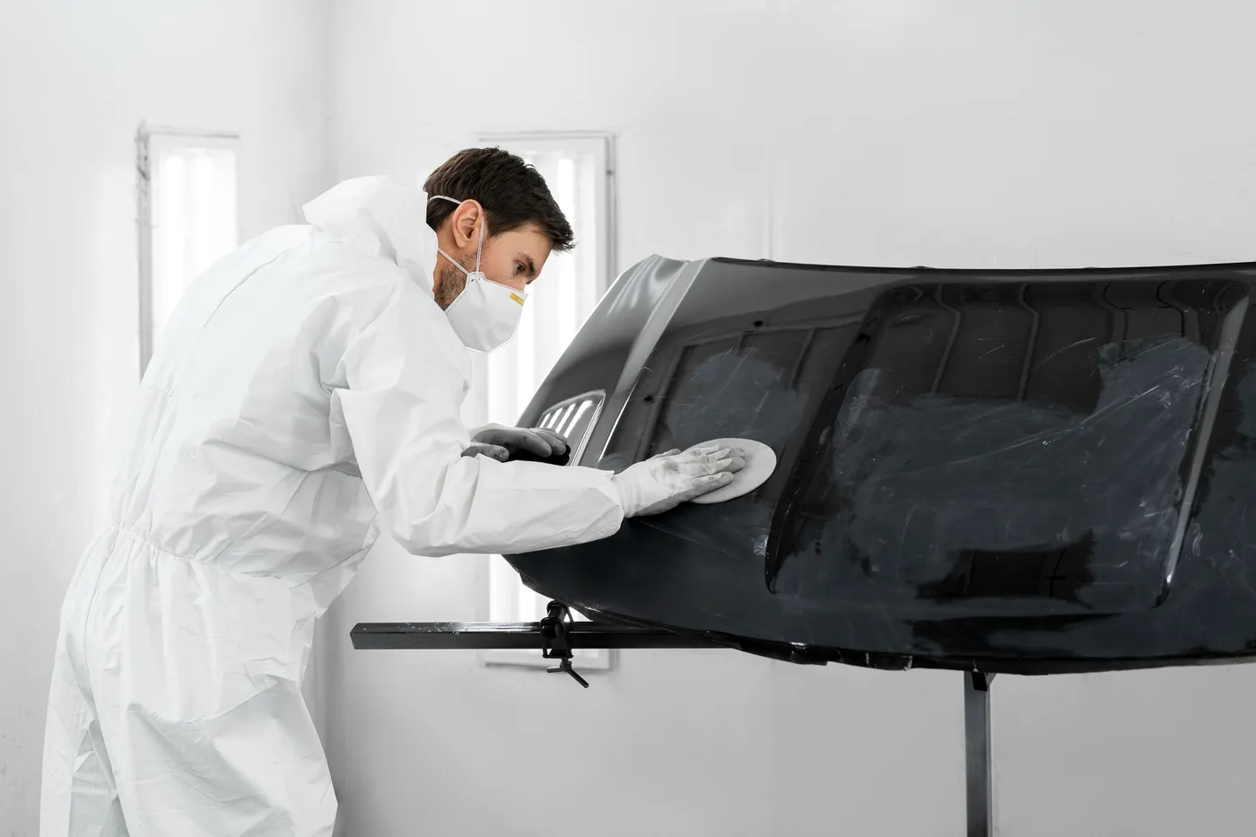 Técnico de carrocería en un taller, limpiando y puliendo un capó de automóvil negro con guantes y mascarilla, en un ambiente controlado y luminoso.
