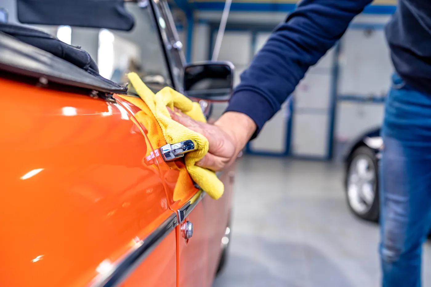 Un hombre limpia con un paño amarillo la puerta de un coche deportivo naranja en un taller de reparación.