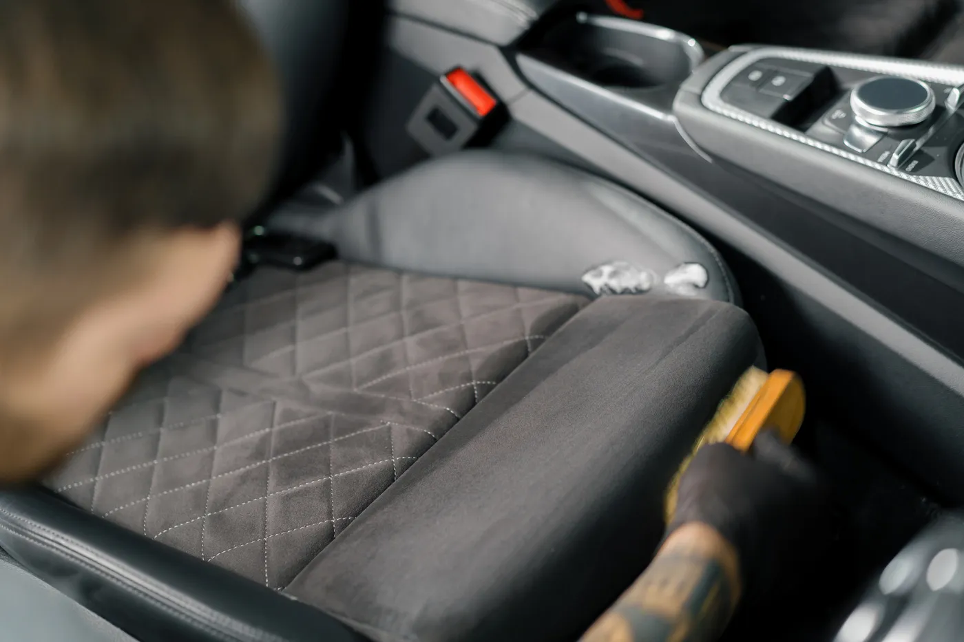 Limpieza de asiento de automóvil con un cepillo, mostrando un enfoque en la tapicería de color gris y detalles de costura.