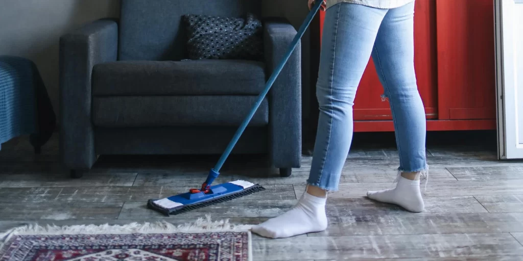 Persona limpiando el suelo de una sala de estar moderna con un trapeador, usando jeans y calcetines blancos, en un ambiente acogedor con un sofá gris y una mesa de centro.