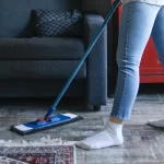 Persona limpiando el suelo de una sala de estar moderna con un trapeador, usando jeans y calcetines blancos, en un ambiente acogedor con un sofá gris y una mesa de centro.