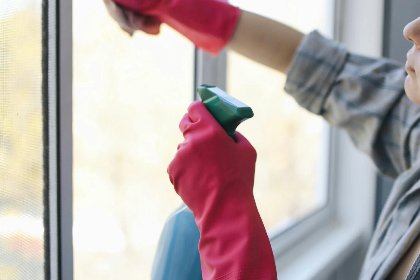 Persona limpiando una ventana con guantes de goma rosas, sosteniendo un spray de limpieza. La imagen muestra un enfoque en la acción de limpiar, resaltando un ambiente luminoso y ordenado.
