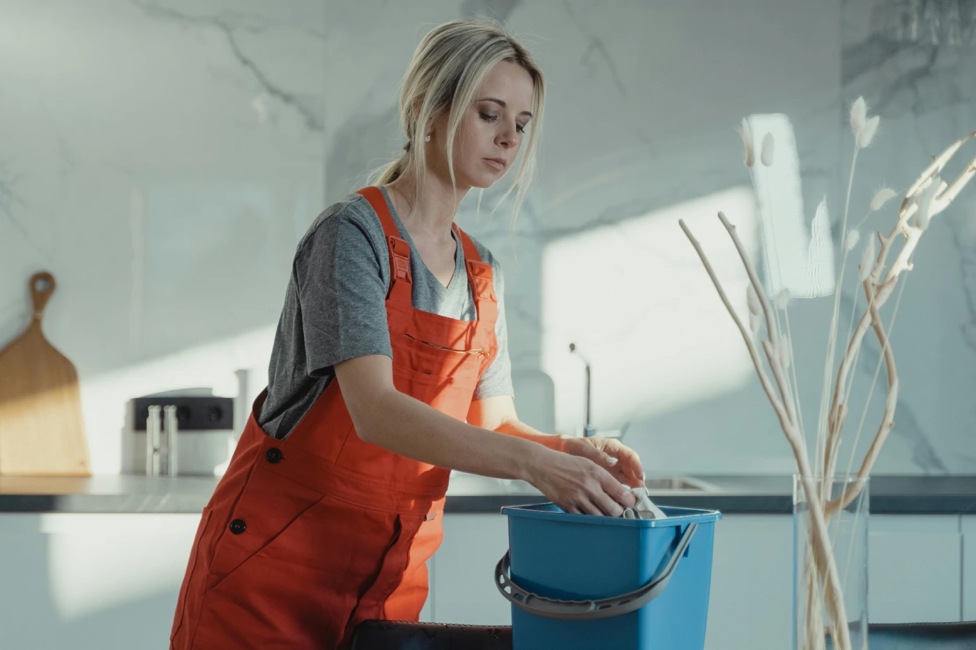 Mujer joven con overol naranja limpiando en la cocina con un balde azul, rodeada de un ambiente moderno y luminoso.
