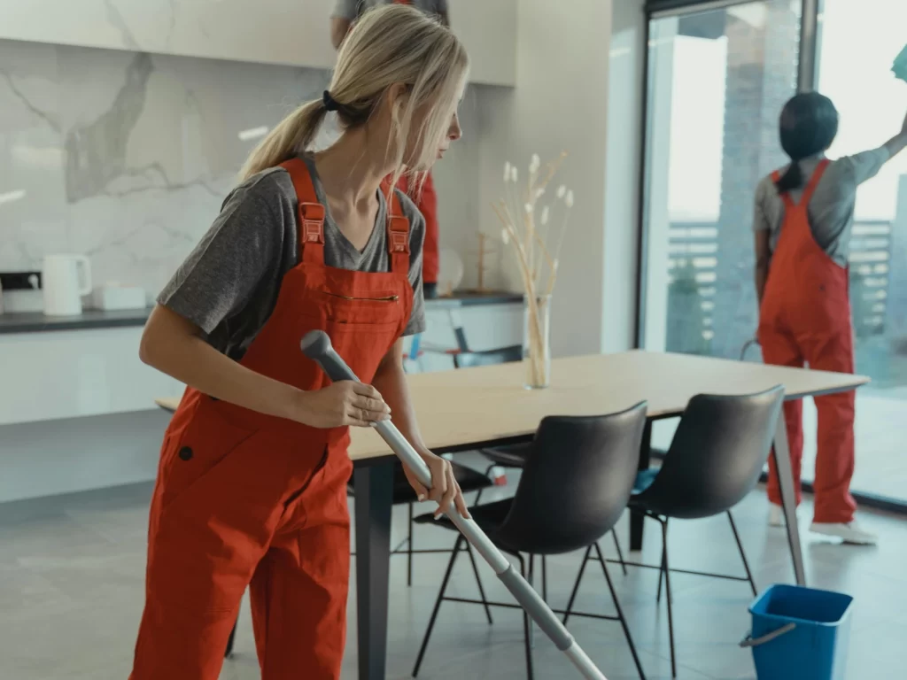 Mujer limpiando el suelo de una cocina moderna con un trapeador, mientras otra persona limpia una ventana. Ambas llevan overoles rojos en un ambiente luminoso y ordenado.