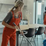 Mujer limpiando el suelo de una cocina moderna con un trapeador, mientras otra persona limpia una ventana. Ambas llevan overoles rojos en un ambiente luminoso y ordenado.